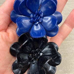 Könnte beinhalten: Eine Brosche mit zwei übereinanderliegenden, mehrblättrigen Blüten aus glänzendem dunkelblauem und schwarzem Kunststoff. Jede Blüte hat in der Mitte eine Ansammlung kleiner, facettierter, farblich passender Perlen, die ein dreidimensionales Design bilden.