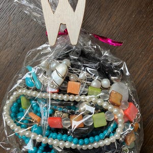 Puede incluir: Una bolsa de pl&aacute;stico transparente llena de una colecci&oacute;n de joyas variadas, incluyendo cuentas turquesas, perlas blancas y cuentas cuadradas de color naranja y verde. La bolsa est&aacute; sellada con un clip de madera clara en forma de letra W.