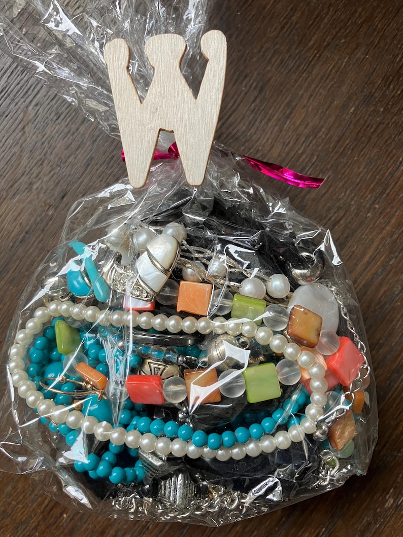 Puede incluir: Una bolsa de pl&aacute;stico transparente llena de una colecci&oacute;n de joyas variadas, incluyendo cuentas turquesas, perlas blancas y cuentas cuadradas de color naranja y verde. La bolsa est&aacute; sellada con un clip de madera clara en forma de letra W.