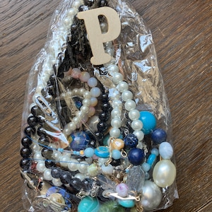 Puede incluir: Una bolsa de pl&aacute;stico transparente llena de una colecci&oacute;n de joyas variadas, incluyendo varios hilos de cuentas en tonos blancos, negros, azules y transl&uacute;cidos, junto con una letra P de madera sujeta a la parte superior con un cordel.