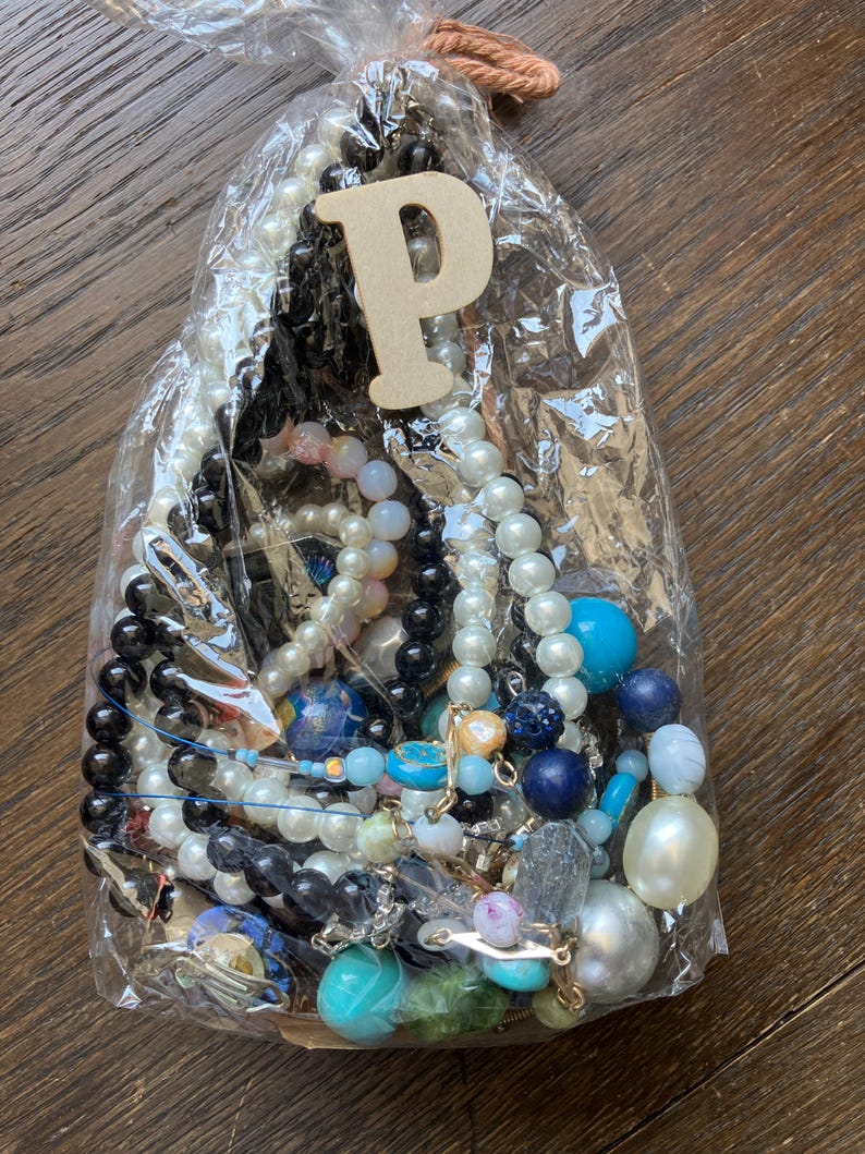 Puede incluir: Una bolsa de pl&aacute;stico transparente llena de una colecci&oacute;n de joyas variadas, incluyendo varios hilos de cuentas en tonos blancos, negros, azules y transl&uacute;cidos, junto con una letra P de madera sujeta a la parte superior con un cordel.