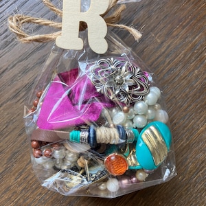 Puede incluir: Una bolsa de pl&aacute;stico transparente llena de joyas variadas que incluyen perlas blancas, una pieza ovalada turquesa y dorada, un broche floral plateado, cuentas marrones y un trozo de tela rosa brillante, atada con una etiqueta de madera con la letra R.
