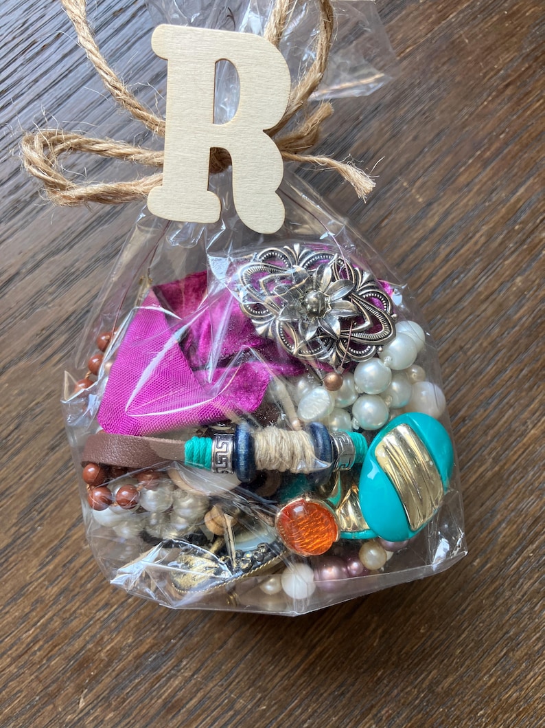 Puede incluir: Una bolsa de pl&aacute;stico transparente llena de joyas variadas que incluyen perlas blancas, una pieza ovalada turquesa y dorada, un broche floral plateado, cuentas marrones y un trozo de tela rosa brillante, atada con una etiqueta de madera con la letra R.