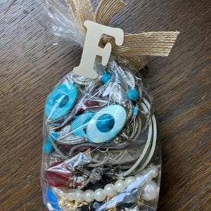 Puede incluir: Una bolsa de pl&aacute;stico transparente llena de joyas variadas que incluyen cuentas ovaladas azul turquesa, perlas blancas artificiales, cadenas de metal plateado y cuentas rojas facetadas. La bolsa est&aacute; atada con una cinta de arpillera y una letra F de madera.
