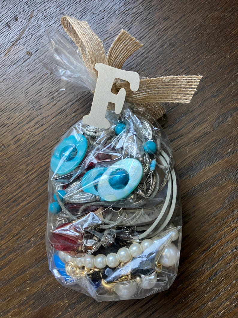 Puede incluir: Una bolsa de pl&aacute;stico transparente llena de joyas variadas que incluyen cuentas ovaladas azul turquesa, perlas blancas artificiales, cadenas de metal plateado y cuentas rojas facetadas. La bolsa est&aacute; atada con una cinta de arpillera y una letra F de madera.