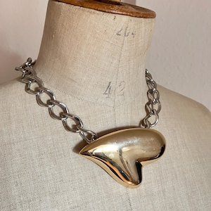 Könnte beinhalten: Eine Halskette mit einem großen, glatten, polierten goldfarbenen Herzanhänger, der an einer dicken, silberfarbenen Metallkette mit ineinandergreifenden ovalen Gliedern hängt. Das Herz hat eine abgerundete, asymmetrische Form mit glänzender Oberfläche.