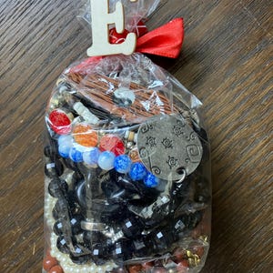 Puede incluir: Una bolsa de pl&aacute;stico transparente llena de joyas variadas que incluyen cuentas negras facetadas, perlas blancas, dijes circulares rojos y naranjas, cuentas azules jaspeadas, un colgante de madera yin-yang y un dije de metal plateado con s&iacute;mbolos grabados.