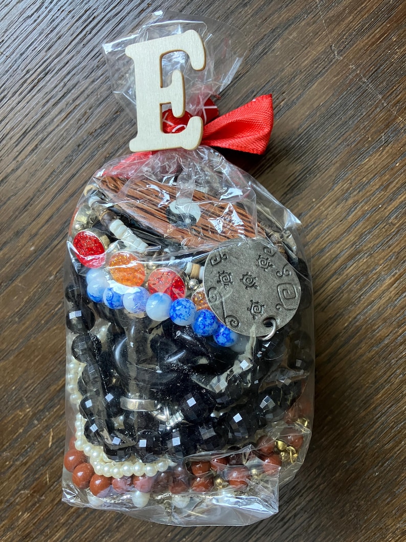 Puede incluir: Una bolsa de pl&aacute;stico transparente llena de joyas variadas que incluyen cuentas negras facetadas, perlas blancas, dijes circulares rojos y naranjas, cuentas azules jaspeadas, un colgante de madera yin-yang y un dije de metal plateado con s&iacute;mbolos grabados.