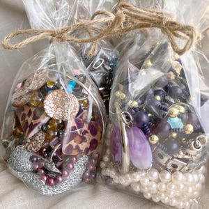 Puede incluir: Dos bolsas de celofán transparente atadas con hilo, llenas de joyas. Una bolsa contiene una mezcla de joyas marrones, doradas y moradas. La otra bolsa tiene joyas moradas, plateadas y blancas, incluyendo perlas y colgantes.