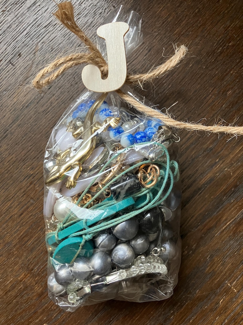 Puede incluir: Una bolsa de pl&aacute;stico transparente llena de joyas variadas que incluyen un dije de lagarto dorado, cuentas jaspeadas azules y blancas, esferas met&aacute;licas plateadas, un cord&oacute;n turquesa y cristales transparentes, atada en la parte superior con cuerda y una letra J de madera.