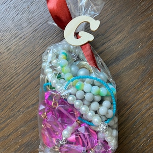 Puede incluir: Una bolsa de pl&aacute;stico transparente llena de bisuter&iacute;a variada, incluyendo perlas blancas de imitaci&oacute;n, cuentas de pl&aacute;stico facetadas de color rosa brillante y un hilo de cuentas turquesa. La bolsa est&aacute; atada con una cinta de raso roja y una letra G de madera.
