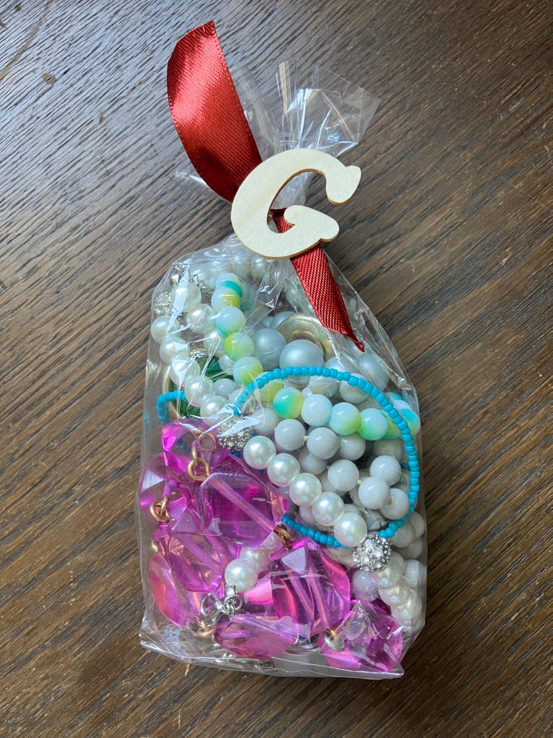 Puede incluir: Una bolsa de pl&aacute;stico transparente llena de bisuter&iacute;a variada, incluyendo perlas blancas de imitaci&oacute;n, cuentas de pl&aacute;stico facetadas de color rosa brillante y un hilo de cuentas turquesa. La bolsa est&aacute; atada con una cinta de raso roja y una letra G de madera.