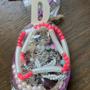 Puede incluir: Una bolsa de pl&aacute;stico transparente que contiene bisuter&iacute;a variada, incluyendo cadenas plateadas, perlas artificiales blancas y un collar de cuentas tubulares de color rosa brillante y blanco. Una letra D de madera est&aacute; sujeta a la parte superior de la bolsa, atada con cordel.