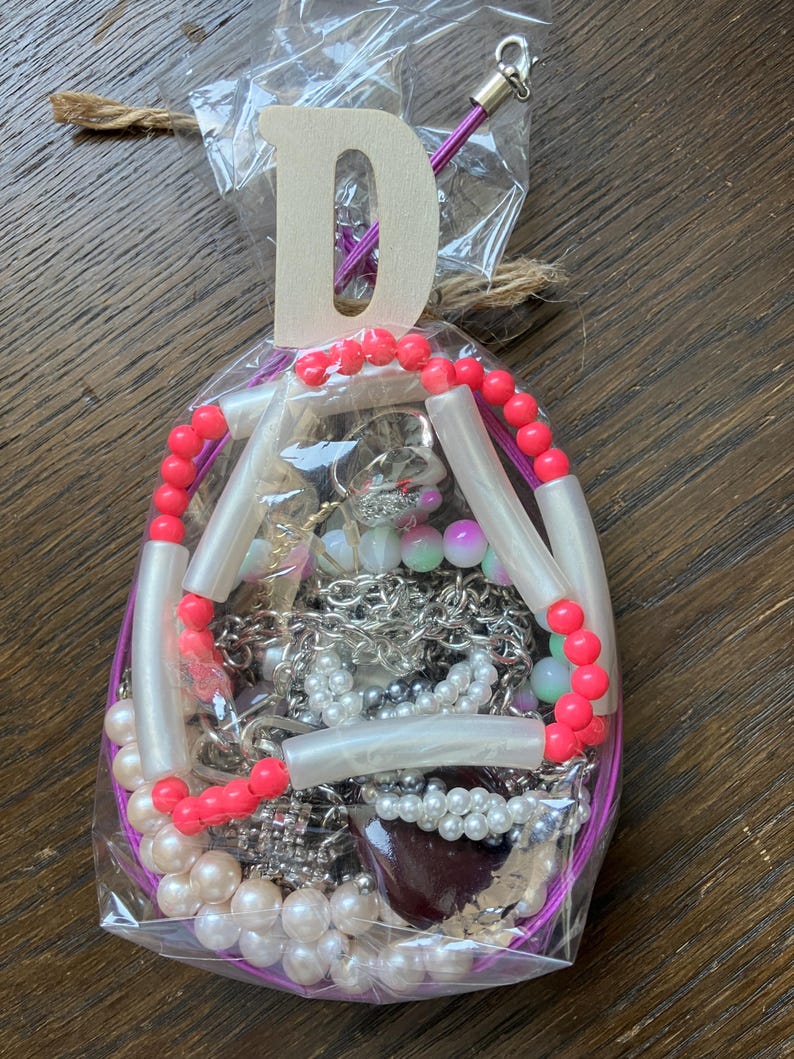 Puede incluir: Una bolsa de pl&aacute;stico transparente que contiene bisuter&iacute;a variada, incluyendo cadenas plateadas, perlas artificiales blancas y un collar de cuentas tubulares de color rosa brillante y blanco. Una letra D de madera est&aacute; sujeta a la parte superior de la bolsa, atada con cordel.