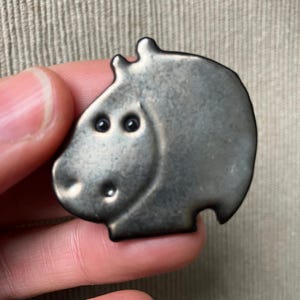 Può includere: Una spilla a forma di ippopotamo color argento. La spilla ha due piccoli occhi rotondi e un piccolo naso rotondo. La testa dell'ippopotamo è leggermente inclinata. La spilla è in metallo e ha una superficie leggermente strutturata.