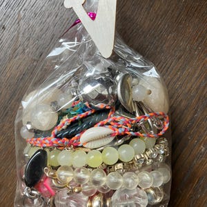 Puede incluir: Una bolsa de pl&aacute;stico transparente llena de un surtido de piezas de joyer&iacute;a, incluyendo pulseras de cuentas, una concha de cauri, esferas met&aacute;licas y cuentas facetadas transparentes, rematada con una etiqueta de madera con la letra V.