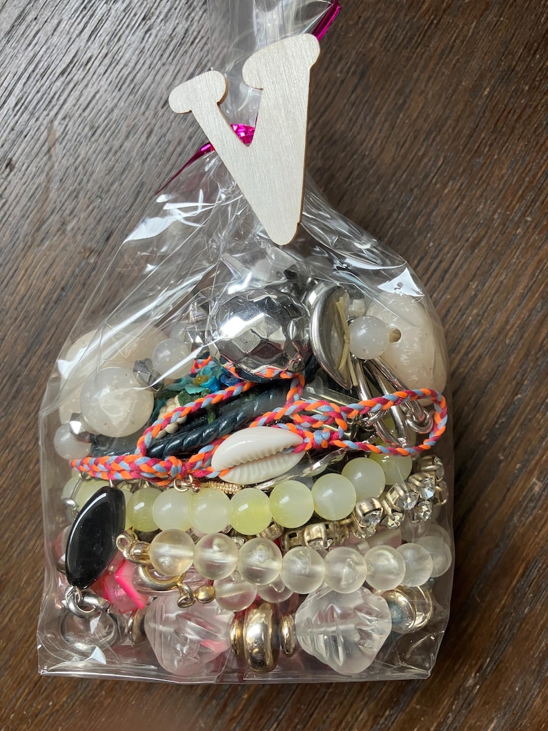 Puede incluir: Una bolsa de pl&aacute;stico transparente llena de un surtido de piezas de joyer&iacute;a, incluyendo pulseras de cuentas, una concha de cauri, esferas met&aacute;licas y cuentas facetadas transparentes, rematada con una etiqueta de madera con la letra V.