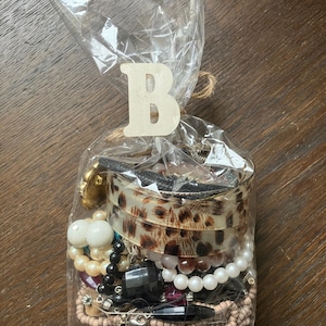 Puede incluir: Una bolsa de pl&aacute;stico transparente llena de joyas variadas, incluyendo un brazalete con estampado de leopardo, cuentas blancas y crema tipo perla, cuentas negras facetadas y hebras de cuentas de semillas color canela. Una letra B de madera est&aacute; unida.