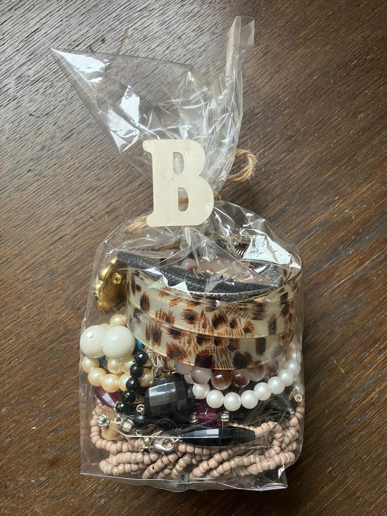 Puede incluir: Una bolsa de pl&aacute;stico transparente llena de joyas variadas, incluyendo un brazalete con estampado de leopardo, cuentas blancas y crema tipo perla, cuentas negras facetadas y hebras de cuentas de semillas color canela. Una letra B de madera est&aacute; unida.