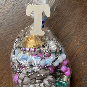 Puede incluir: Una bolsa de pl&aacute;stico transparente llena de bisuter&iacute;a variada, incluyendo piezas en forma de flor blanca, cuentas iridiscentes, cuentas esf&eacute;ricas rosas y azules, y elementos met&aacute;licos. Una letra T de madera clara est&aacute; sujeta a la parte superior.