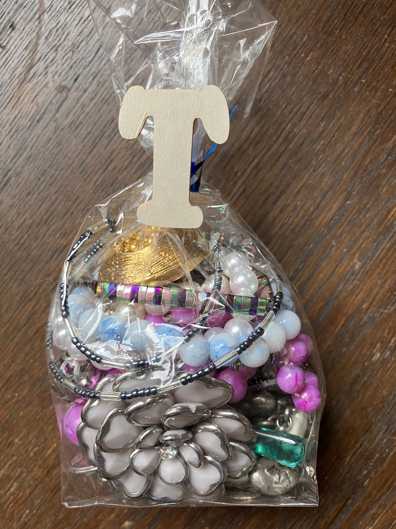 Puede incluir: Una bolsa de pl&aacute;stico transparente llena de bisuter&iacute;a variada, incluyendo piezas en forma de flor blanca, cuentas iridiscentes, cuentas esf&eacute;ricas rosas y azules, y elementos met&aacute;licos. Una letra T de madera clara est&aacute; sujeta a la parte superior.