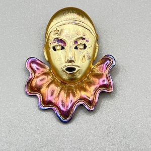 Könnte beinhalten: Goldfarbene Metallbrosche in Form eines Narrenkopfes. Der Narr hat einen kleinen Stern auf dem Kopf, mit einem tropfenförmigen Detail auf der Wange. Der Rüschenkragen hat schillernde rosa, goldene und blaue Farbtöne.