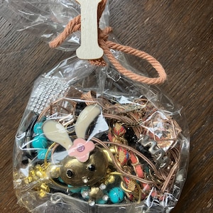 Puede incluir: Una bolsa de pl&aacute;stico transparente llena de bisuter&iacute;a variada, incluyendo un dije de conejo dorado con orejas blancas y una flor rosa, cuentas turquesas y cadenas met&aacute;licas. La bolsa est&aacute; atada con un cord&oacute;n color melocot&oacute;n y una etiqueta de madera con la letra I.