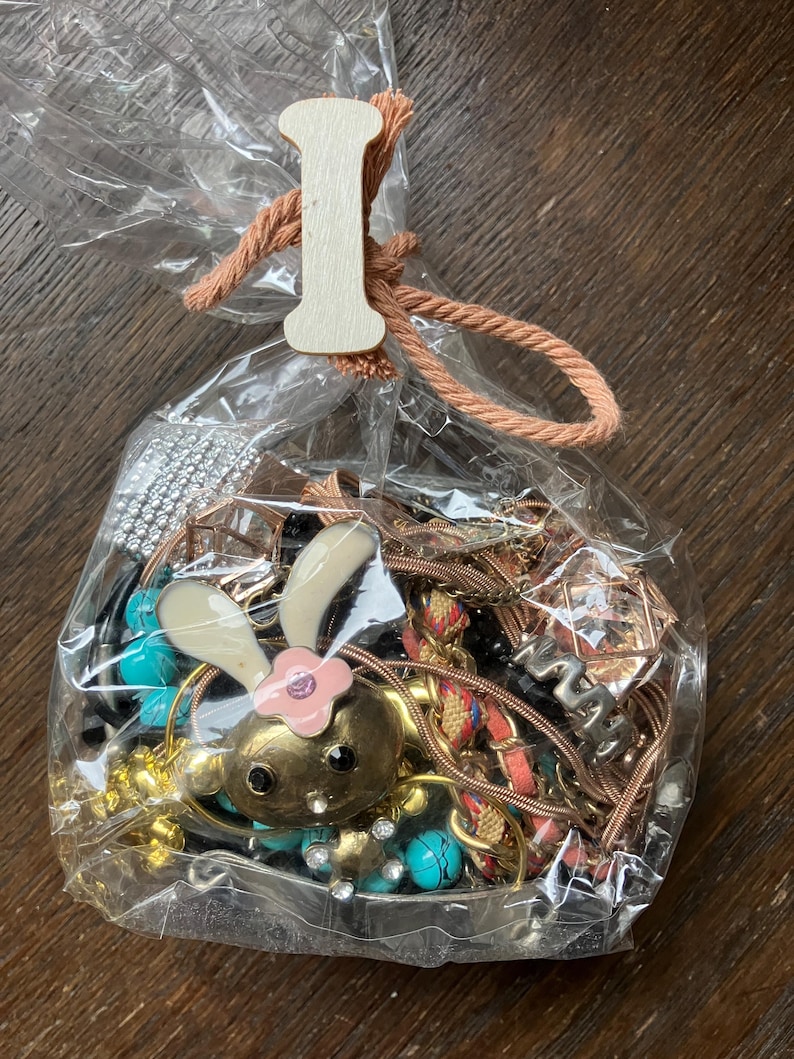 Puede incluir: Una bolsa de pl&aacute;stico transparente llena de bisuter&iacute;a variada, incluyendo un dije de conejo dorado con orejas blancas y una flor rosa, cuentas turquesas y cadenas met&aacute;licas. La bolsa est&aacute; atada con un cord&oacute;n color melocot&oacute;n y una etiqueta de madera con la letra I.