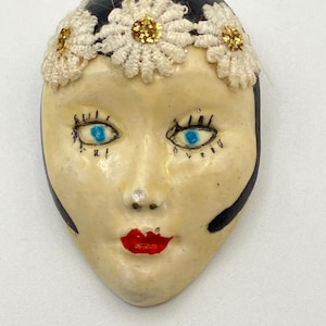 Könnte beinhalten: Eine dekorative Brosche in Form eines menschlichen Gesichts mit cremefarbenem Teint, leuchtend blauen Augen und roten Lippen. Das Gesicht ist von dunklem Haar umrahmt, das mit drei weißen, gehäkelten Gänseblümchen mit goldenen Glitzerzentren verziert ist.