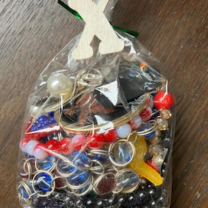 Puede incluir: Una bolsa de pl&aacute;stico transparente llena de una variedad de joyas coloridas, incluyendo cuentas azules, rojas y negras, cadenas met&aacute;licas y una cuenta blanca similar a una perla. Una letra X de madera clara est&aacute; sujeta a la parte superior.
