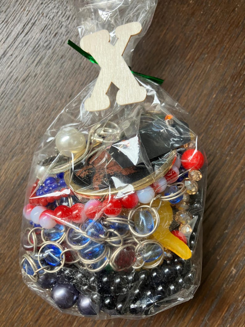 Puede incluir: Una bolsa de pl&aacute;stico transparente llena de una variedad de joyas coloridas, incluyendo cuentas azules, rojas y negras, cadenas met&aacute;licas y una cuenta blanca similar a una perla. Una letra X de madera clara est&aacute; sujeta a la parte superior.