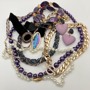 Könnte beinhalten: Eine Sammlung verschiedener Schmuckstücke, darunter Halsketten aus violetten und weißen Perlen, eine grobe goldfarbene Gliederkette mit Knebelverschluss, ein schwarzes Samt-Halsband, zwei lavendelfarbene herzförmige Ohrringe und ein Armband.