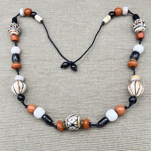 Könnte beinhalten: Eine handgefertigte Halskette mit einer Vielzahl von Perlen in Orange, Weiß, Schwarz und Creme. Die Perlen sind auf einer schwarzen Kordel aufgefädelt, was ein einzigartiges Accessoire ergibt. Die Halskette hat eine Tribal- oder Boho-Ästhetik. Die Halskette ist ca. 45 cm lang.