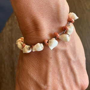Puede incluir: Una pulsera que consiste en un cordón marrón ensartado con pequeñas conchas blancas y cuentas redondas y texturizadas de color marrón rojizo. Las conchas están dispuestas en un patrón repetitivo con las cuentas, creando un diseño circular.