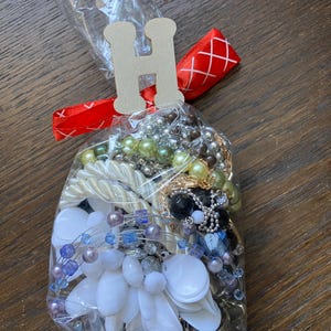 Puede incluir: Una bolsa de pl&aacute;stico transparente llena de una colecci&oacute;n de joyas variadas, que incluye cuentas blancas, azules, verdes y negras, cadenas met&aacute;licas y piezas blancas con forma de flor. La bolsa est&aacute; atada con una cinta roja y una etiqueta de madera con la letra H.