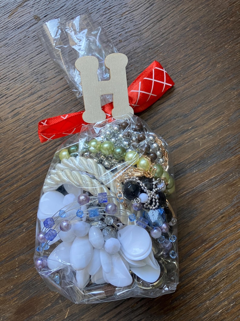 Puede incluir: Una bolsa de pl&aacute;stico transparente llena de una colecci&oacute;n de joyas variadas, que incluye cuentas blancas, azules, verdes y negras, cadenas met&aacute;licas y piezas blancas con forma de flor. La bolsa est&aacute; atada con una cinta roja y una etiqueta de madera con la letra H.