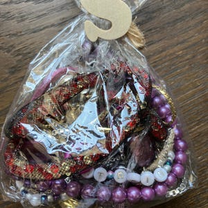 Puede incluir: Una bolsa de pl&aacute;stico transparente que contiene una colecci&oacute;n de joyas variadas, incluyendo cuentas moradas tipo perla, una pulsera de malla roja y negra, y cuentas blancas con letras que forman TRIN. Una letra S de madera est&aacute; atada con cuerda.