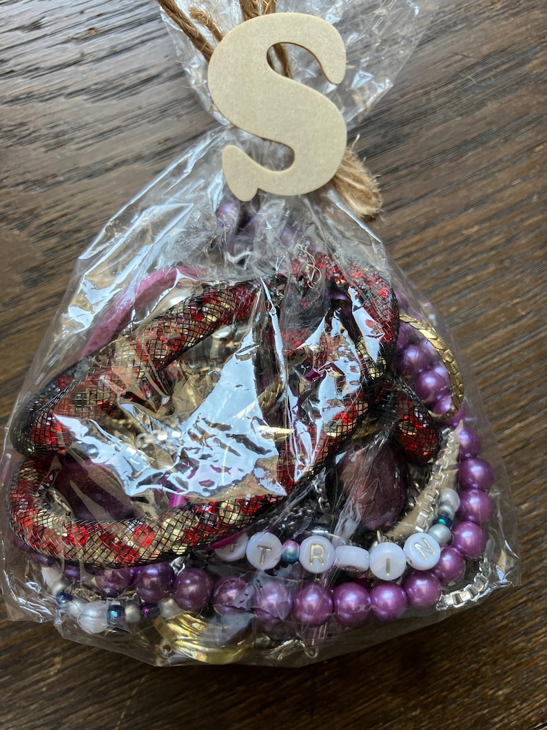 Puede incluir: Una bolsa de pl&aacute;stico transparente que contiene una colecci&oacute;n de joyas variadas, incluyendo cuentas moradas tipo perla, una pulsera de malla roja y negra, y cuentas blancas con letras que forman TRIN. Una letra S de madera est&aacute; atada con cuerda.