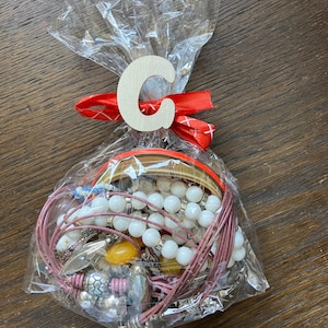 Puede incluir: Una colecci&oacute;n de pulseras y piezas de joyer&iacute;a variadas empaquetadas en una bolsa de pl&aacute;stico transparente. La bolsa est&aacute; atada con una cinta roja y presenta una letra C de madera de color claro adherida al frente. La joyer&iacute;a incluye cuentas blancas, bandas met&aacute;licas y cordones rosas.