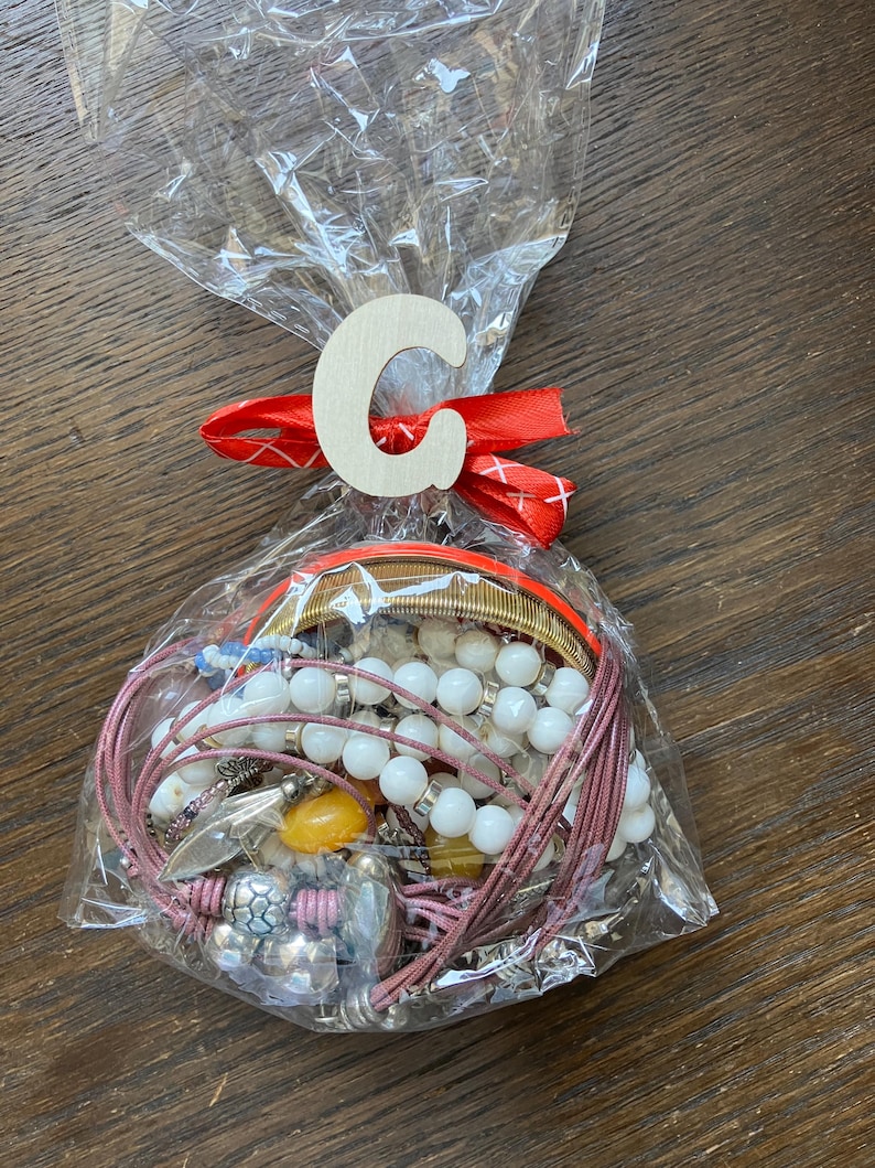 Puede incluir: Una colecci&oacute;n de pulseras y piezas de joyer&iacute;a variadas empaquetadas en una bolsa de pl&aacute;stico transparente. La bolsa est&aacute; atada con una cinta roja y presenta una letra C de madera de color claro adherida al frente. La joyer&iacute;a incluye cuentas blancas, bandas met&aacute;licas y cordones rosas.