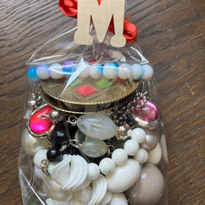 Puede incluir: Una bolsa de pl&aacute;stico transparente llena de un surtido de cuentas para manualidades y componentes de joyer&iacute;a, incluyendo esferas blancas, negras, rosas e iridiscentes, un dije circular color bronce y una etiqueta de madera con la letra M.
