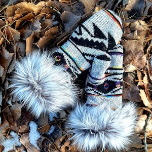 Może przedstawiać: Para dzianinowych rękawic z czarno-białym geometrycznym wzorem i puszystym, szarym futrzanym wykończeniem. Rękawice leżą na posłaniu z brązowych liści z płatami śniegu, sugerując zimowy motyw.