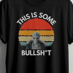 Pode incluir: Camiseta preta com um gráfico de pôr do sol retrô e uma figura alienígena. O texto "This is some BULLSH*T" está impresso na camiseta.