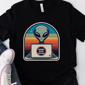 以下が含まれることがあります： 夕焼けの背景に、ラップトップを使用する宇宙人のレトロなグラフィックが描かれた黒いTシャツ。グラフィックには「Works on my machine」というテキストがあります。