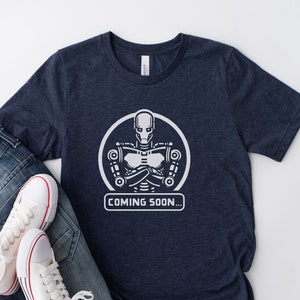 Puede incluir: Camiseta azul marino con un diseño de robot blanco dentro de un círculo y el texto "COMING SOON...". La camiseta se muestra con vaqueros azules y zapatillas blancas con rayas rojas.