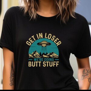 May include: Black t-shirt with a retro graphic featuring a UFO over a mountain range. The text reads "GET IN LOSER WE'RE DOING BUTT STUFF" in a vintage style font. The design is in teal, beige, and black.