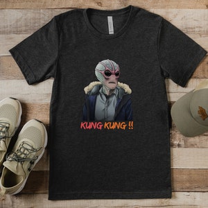 May include: A dark gray t-shirt with a graphic of a creature with pink skin and large eyes wearing a blue jacket. The text "KUNG KUNG!!" is printed in orange below the graphic.