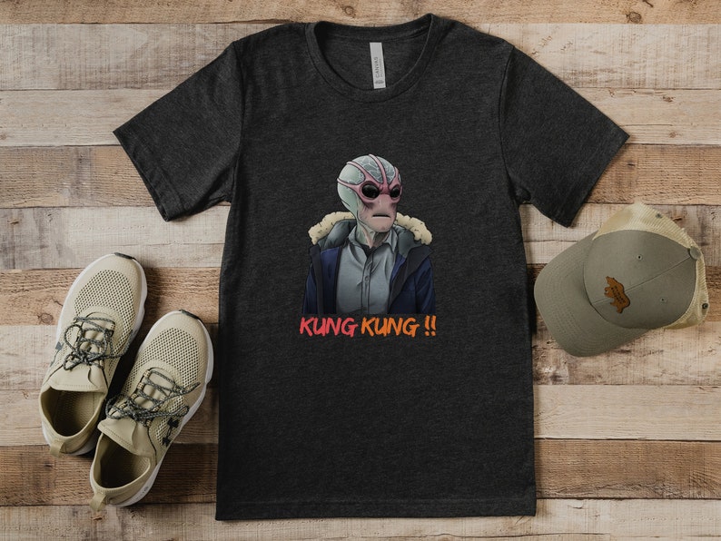 May include: A dark gray t-shirt with a graphic of a creature with pink skin and large eyes wearing a blue jacket. The text "KUNG KUNG!!" is printed in orange below the graphic.