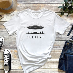 Könnte beinhalten: Weißes T-Shirt mit einem schwarzen Aufdruck eines UFOs, das über ein Gebirge mit Bäumen fliegt, und dem Wort "BELIEVE" darunter. Das Shirt liegt flach auf einer Holzoberfläche mit einem Strohhut, einer Brille, einer Uhr und einer Jeans.
