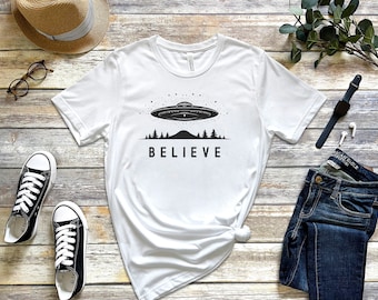 Puede incluir: Camiseta blanca con un gráfico negro de un OVNI volando sobre una cordillera con árboles y la palabra "BELIEVE" debajo. La camiseta está colocada sobre una superficie de madera con un sombrero de paja, gafas, un reloj y unos vaqueros.
